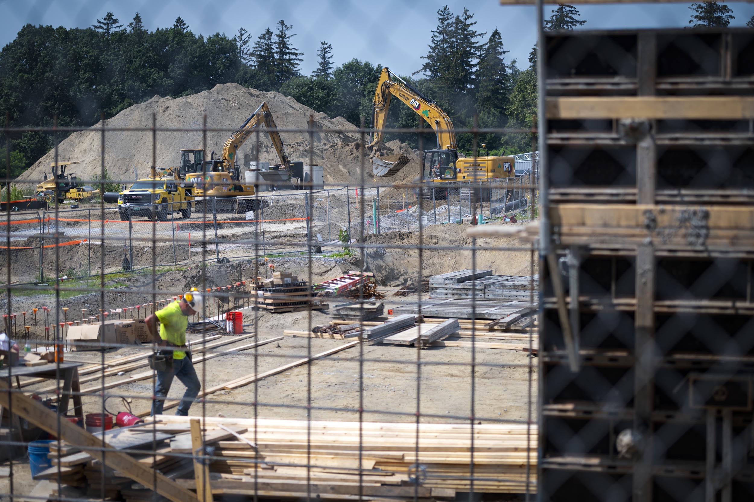 Construction site with excavators moving dirt, a worker carrying materials, and stacks of wood and equipment scattered around, with trees and sand piles in the background.