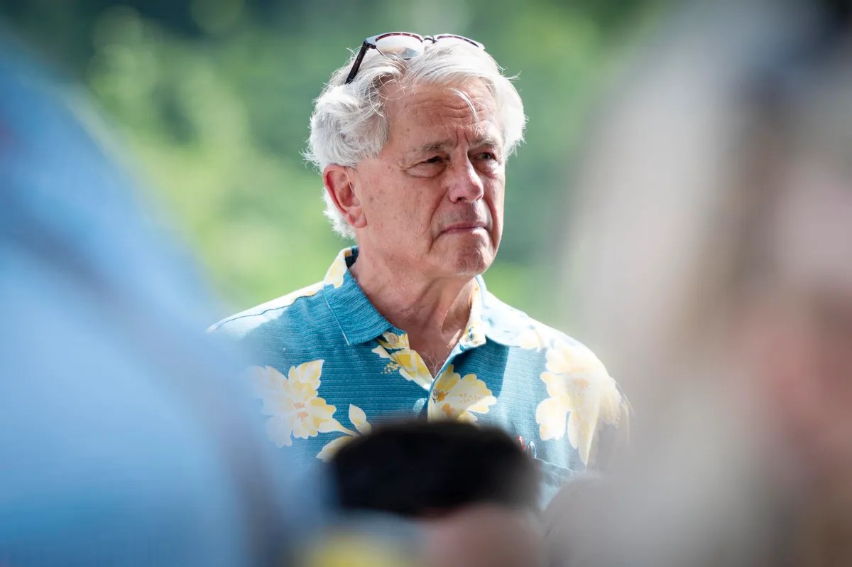 Man with white hair and glasses on his head wearing a blue shirt with yellow flowers, standing outdoors with a blurred background.