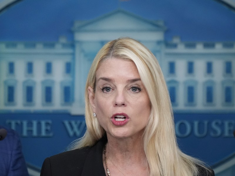 A woman with long blonde hair speaks at a podium with microphones, with the White House emblem visible in the background.