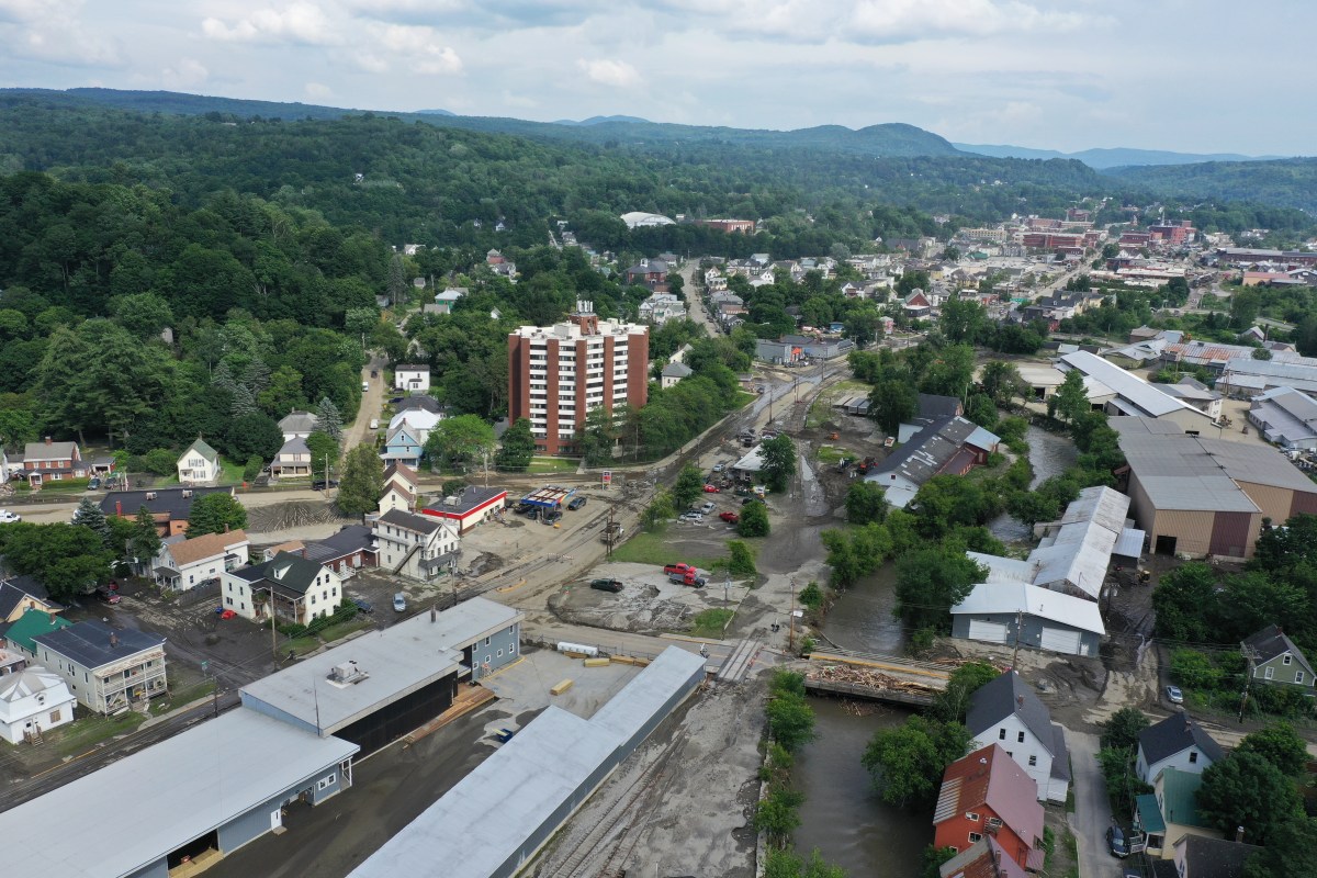 Aerial view of a small town with a mix of residential and industrial buildings, muddy roads, and visible flooding along a riverbank.