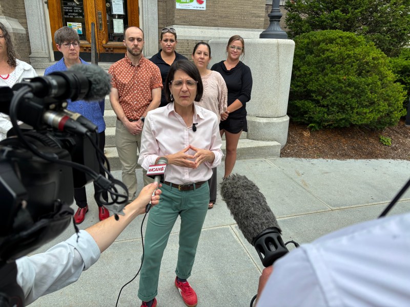 A person speaks to reporters outdoors as several people stand behind her; microphones and cameras are visible in the foreground.