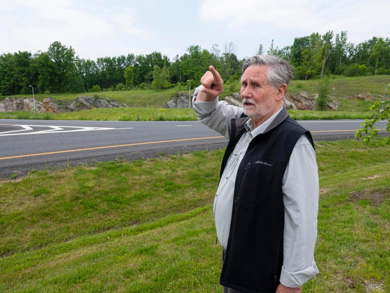 An older man standing on grass near a road points ahead with his right hand. Trees and rocky terrain are visible in the background under a partly cloudy sky.