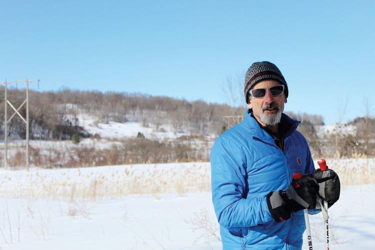 A man in a blue jacket and beanie holds ski poles, standing in a snowy landscape with trees and power lines in the background.