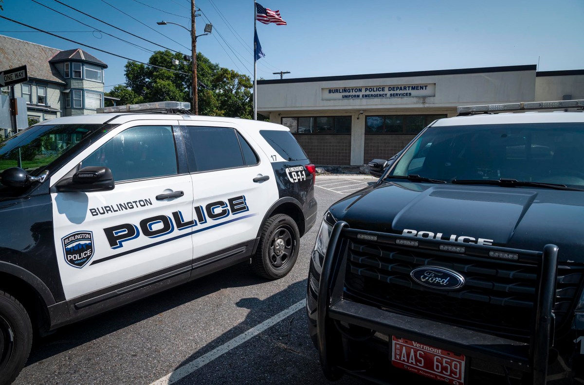 Two police SUVs are parked in front of the Burlington Police Department building, which has American and state flags flying above it.