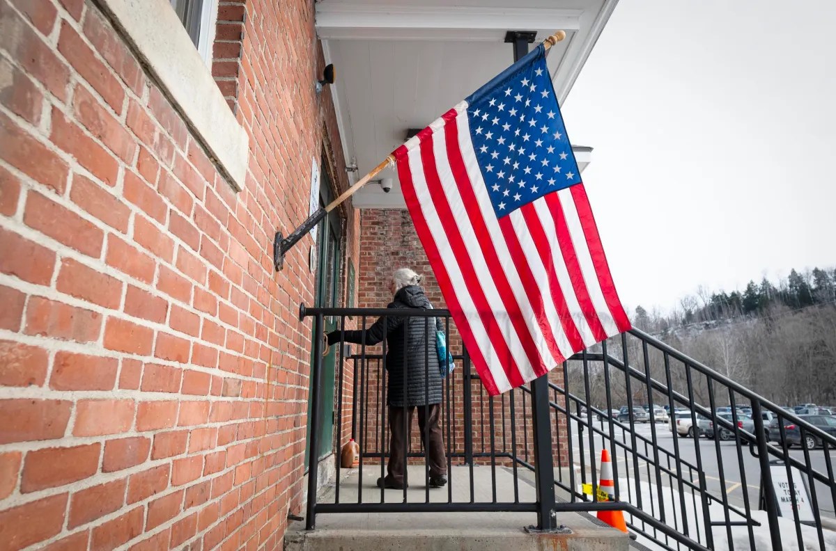 Person standing at an entrance on a brick building with a large American flag displayed.