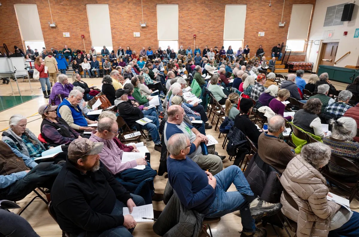 Crowd of people seated in a large room with brick walls, facing forward. Some individuals hold papers, while others watch from the back.