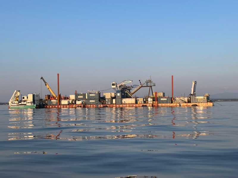 A large barge equipped with various cranes, machinery, and containers floats on calm water under a clear sky.