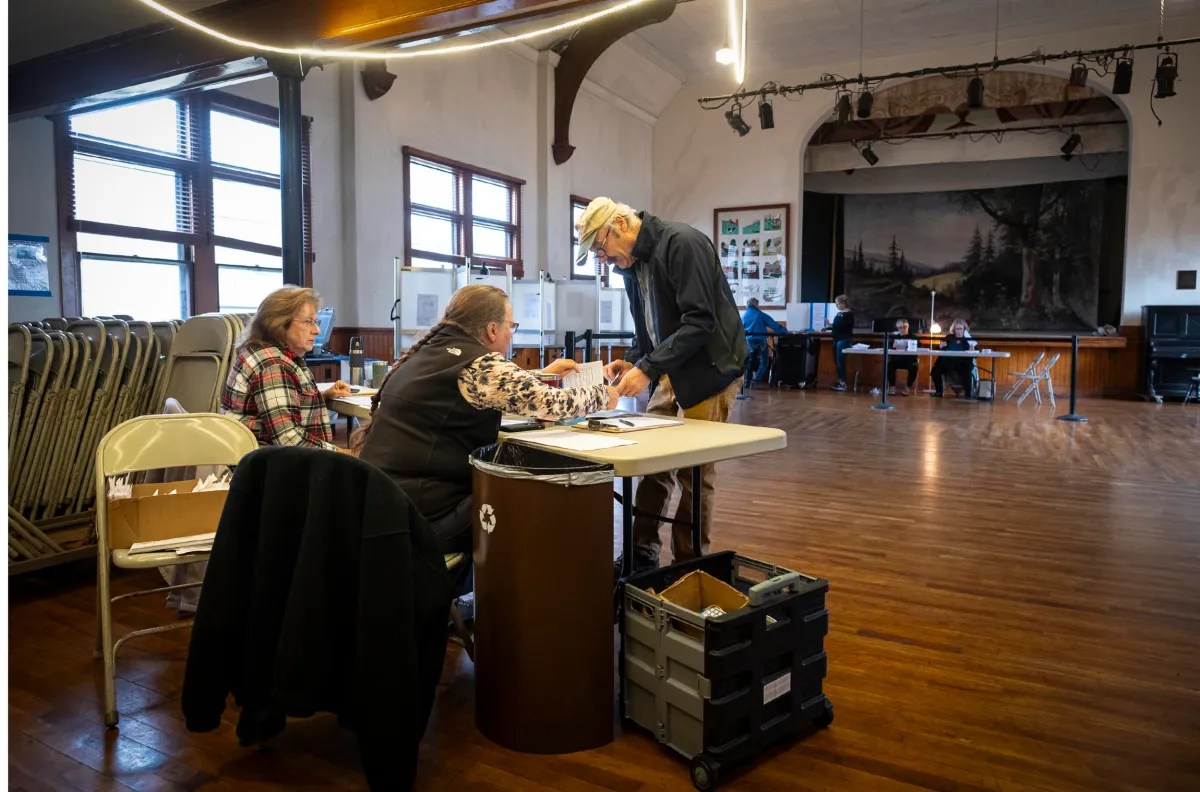 A man signs a document at a table in a large room with two women seated. Voting booths and chairs are visible in the background.