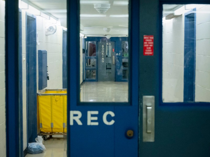A view through a glass door marked "REC" into a hallway with a yellow cleaning cart, white walls, and another door labeled "BOOKING.
