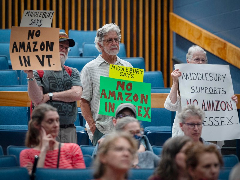 People at a meeting hold signs reading "No Amazon in VT" and "No Amazon in Essex" in protest, while others sit quietly in a blue-seated auditorium.