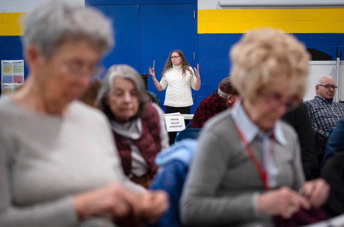 A woman stands in front of a blue door, gesturing with her hands. Several seated people are blurred in the foreground, focusing on various activities.