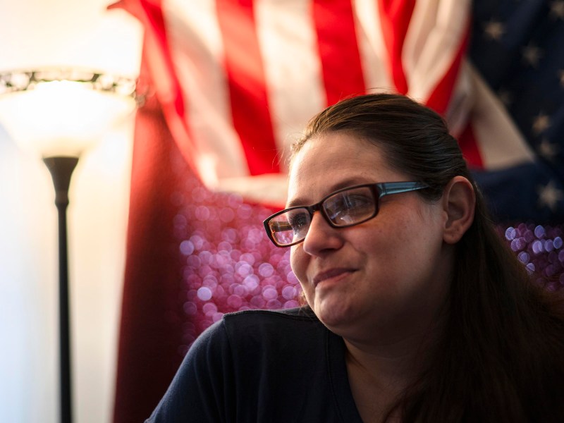 Woman with glasses sits indoors in front of an American flag and a lit floor lamp, with a reflective, red background behind her.