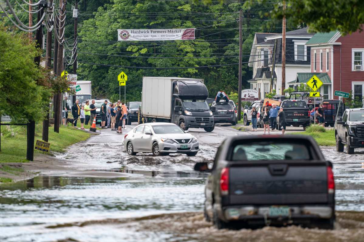 People stand on a flooded street with vehicles attempting to navigate through the water. A sign for a farmers market and various buildings can be seen in the background.