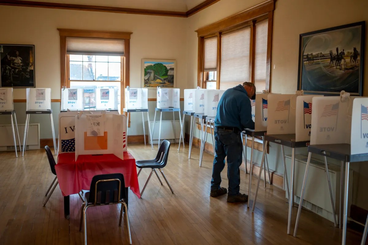 A person votes in a room with multiple voting booths and American flags. One table with chairs is in the center.