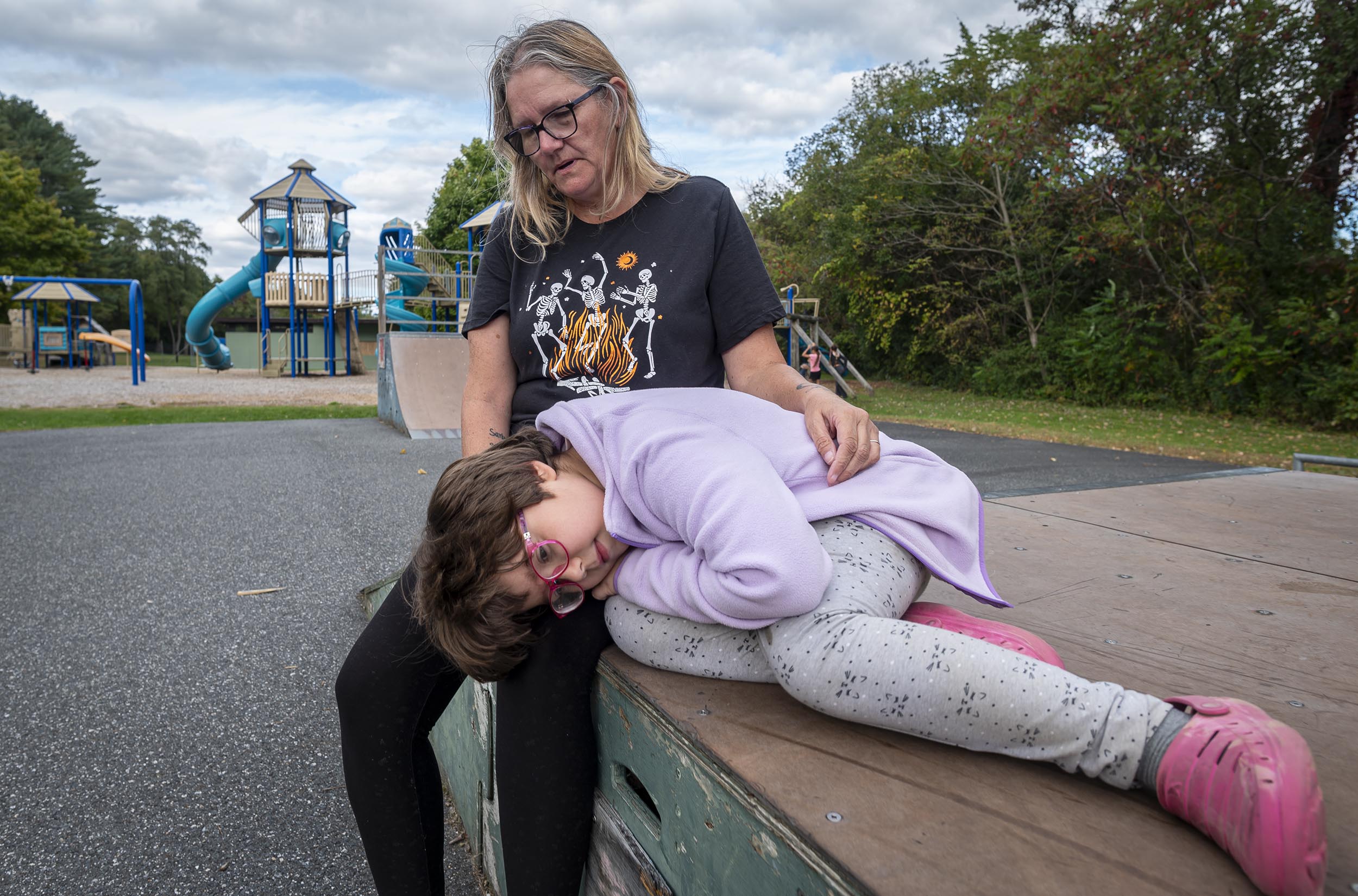 An adult holds a child who is lying down on a platform at a playground with slides visible in the background. The child appears to be resting. Both are outdoors on a cloudy day.