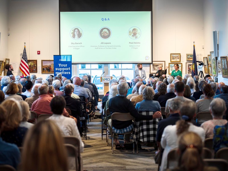 A full audience attends a presentation in a large room featuring a Q&A panel discussion. Three speakers are shown on a projection screen at the front, with an American flag and framed pictures on the walls.
