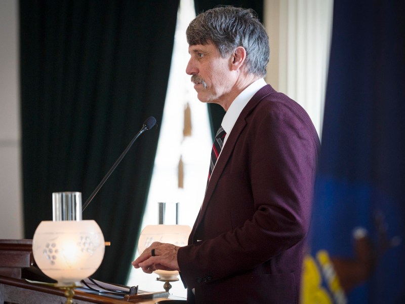 A man in a suit speaks at a podium with a microphone, standing next to a flag and lamps, in a formal setting.