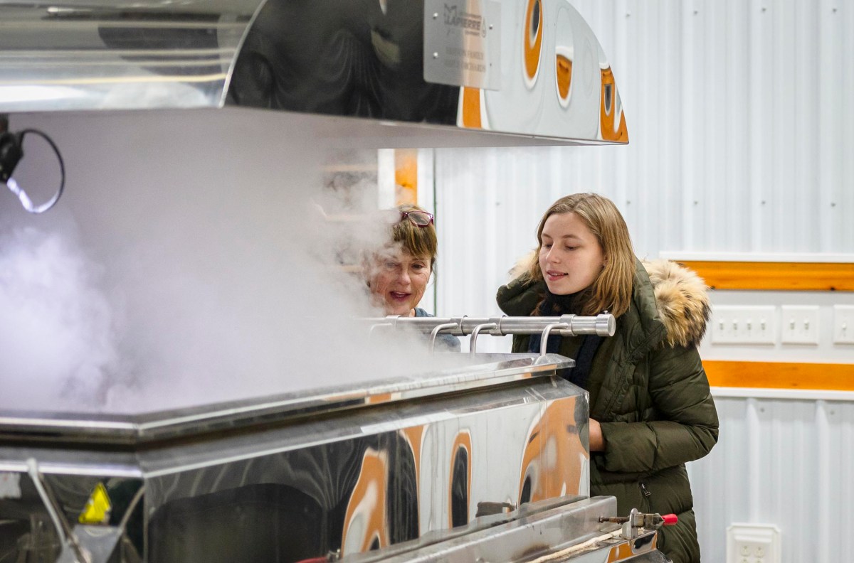 Two people observe a machine producing steam or vapor inside an industrial setting. Both wear warm coats.