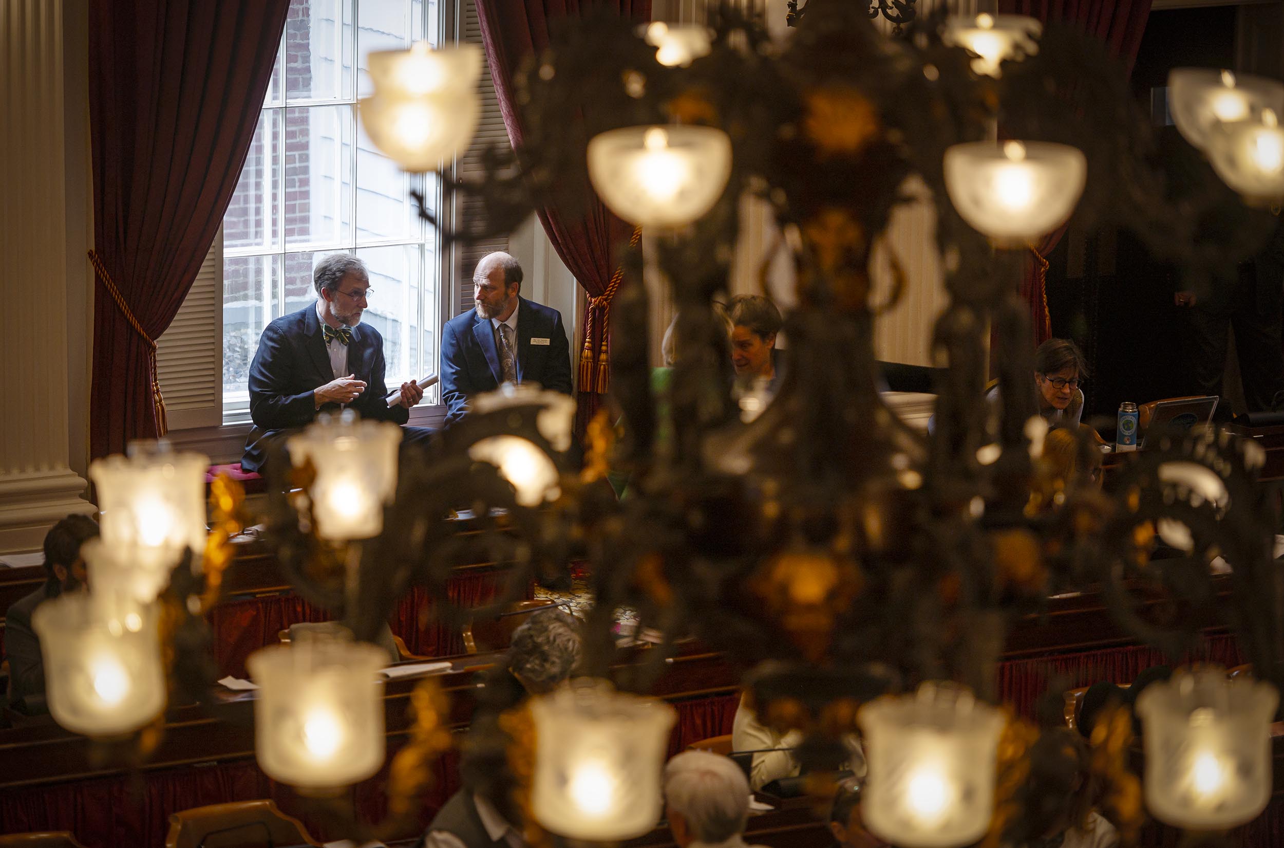 Two men conversing in a grand, dimly lit hall with large windows and ornate metal candelabras emitting soft light foreground.