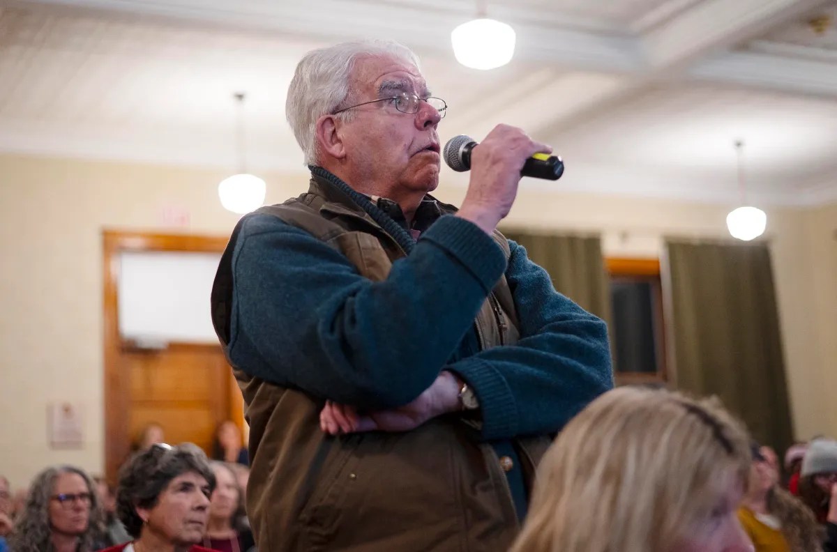 An older man stands in a crowded room, holding a microphone and speaking.