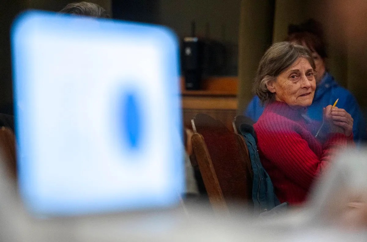 An older woman in a red sweater sits in a chair, holding a pencil and looking intently to the side. A blurred screen is visible in the foreground.