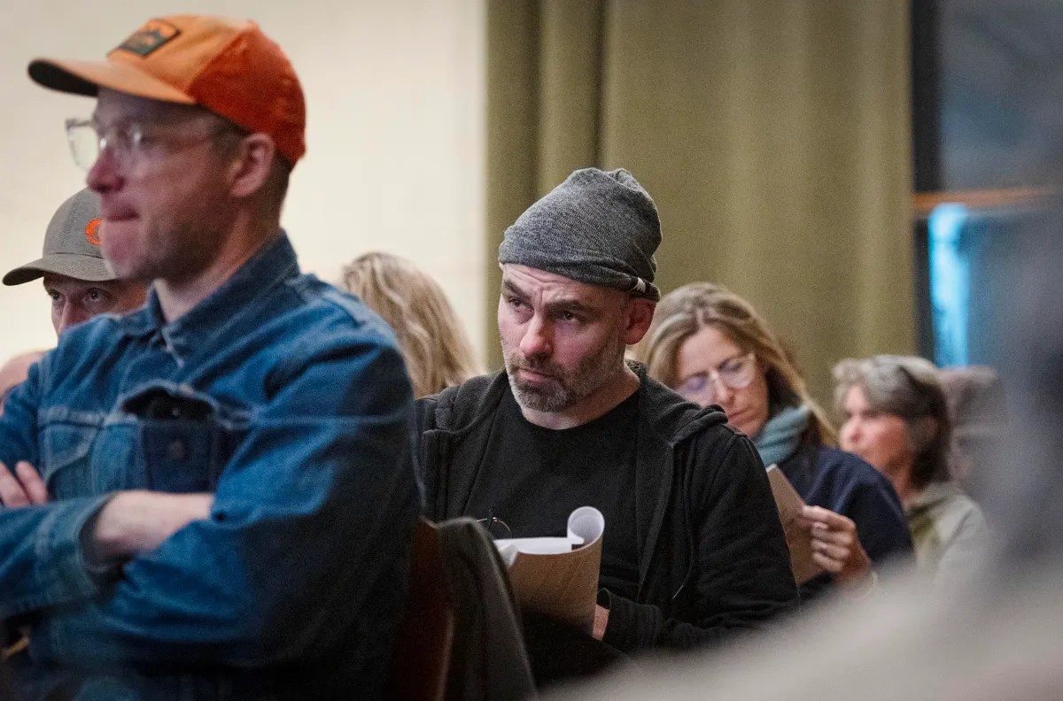 People sitting attentively in a room, holding papers. A man in a gray beanie is prominently in focus among the group.