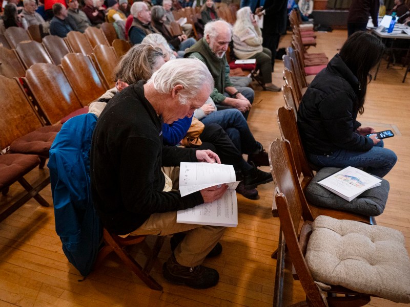 A group of people seated in a hall, some reading documents, others on phones. Wooden chairs and a wooden floor are visible.