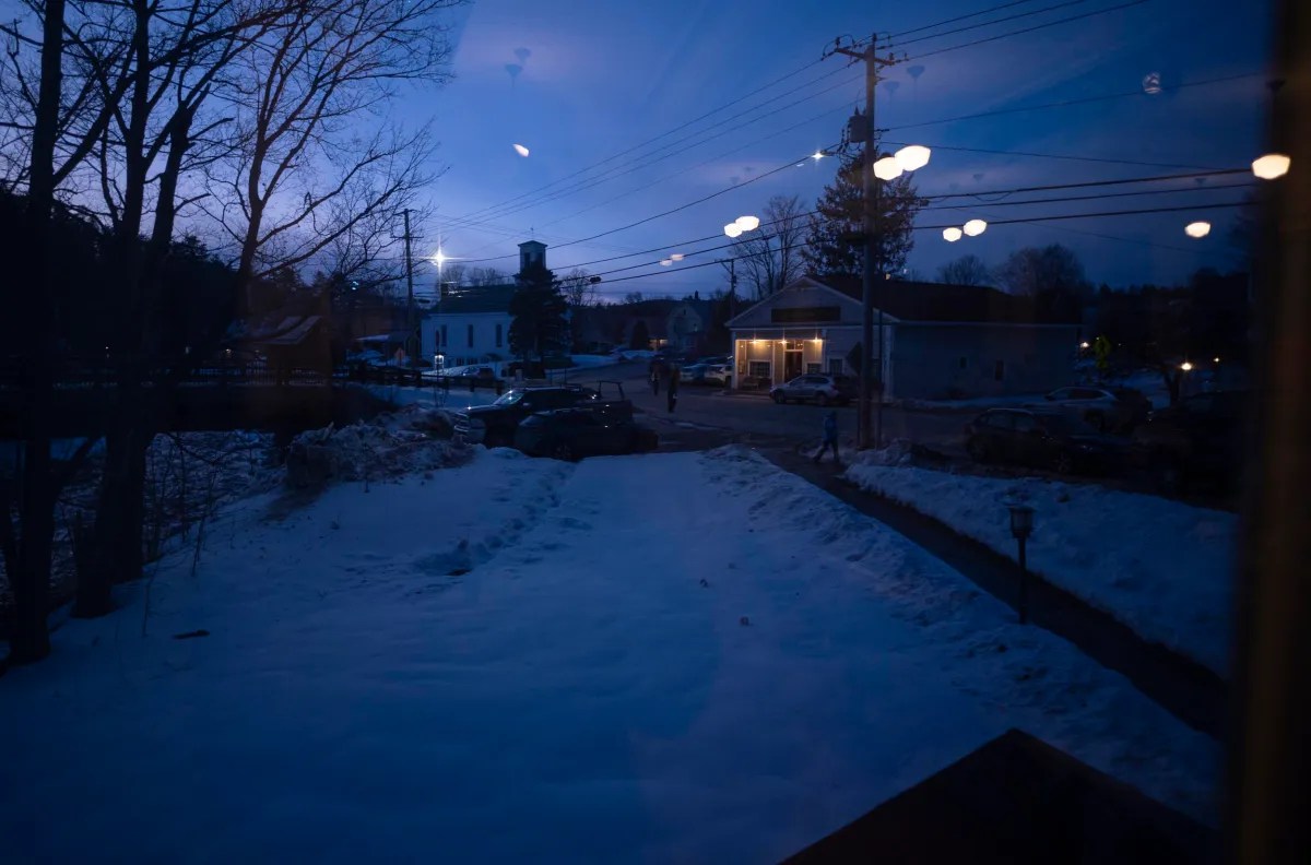 Evening scene of a snowy street with parked cars and buildings. Streetlights are on, and reflections of indoor lights are visible in the window.