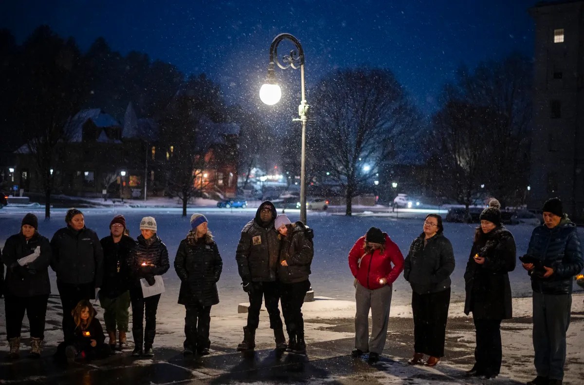 A group of people stands under a streetlight at night in a snowy setting, some holding candles.