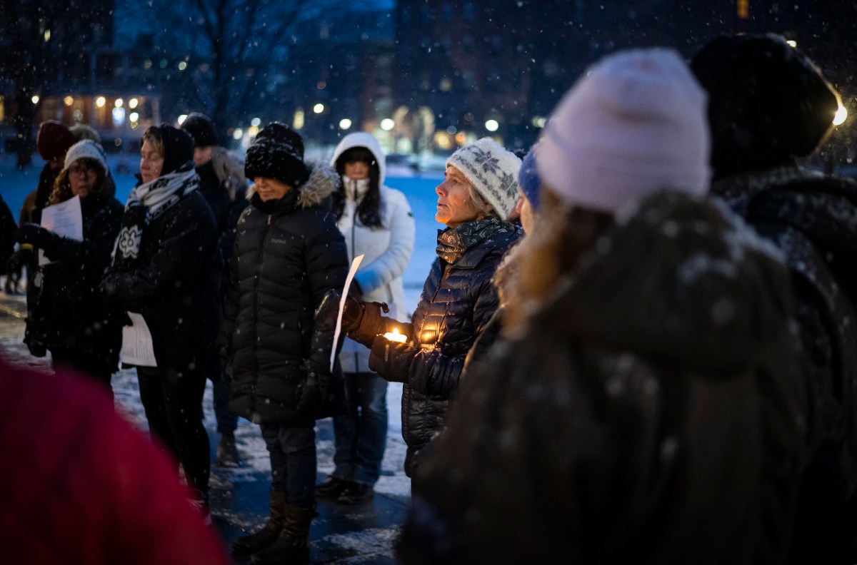 A group of people in winter clothing participate in a nighttime candlelight vigil in a snowy outdoor setting.