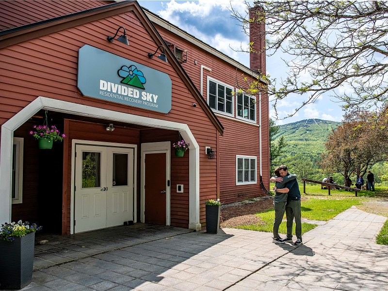 Two people hug outside the entrance of a red building labeled "Divided Sky Residential Recovery Program," with greenery and mountains visible in the background.