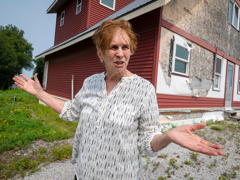 An older woman stands outside a red house with damaged siding, gesturing with both arms outstretched on a gravel and grass area.