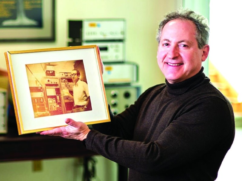 Man in a black turtleneck smiles and holds a framed photo of another man in a control room with equipment.