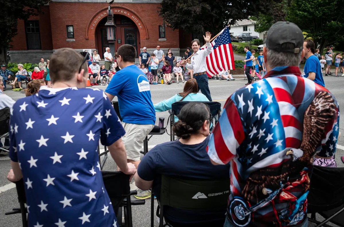 A crowd of people, many wearing American flag-themed clothing, gather outdoors as a man holding an American flag waves and smiles. A brick building is visible in the background.