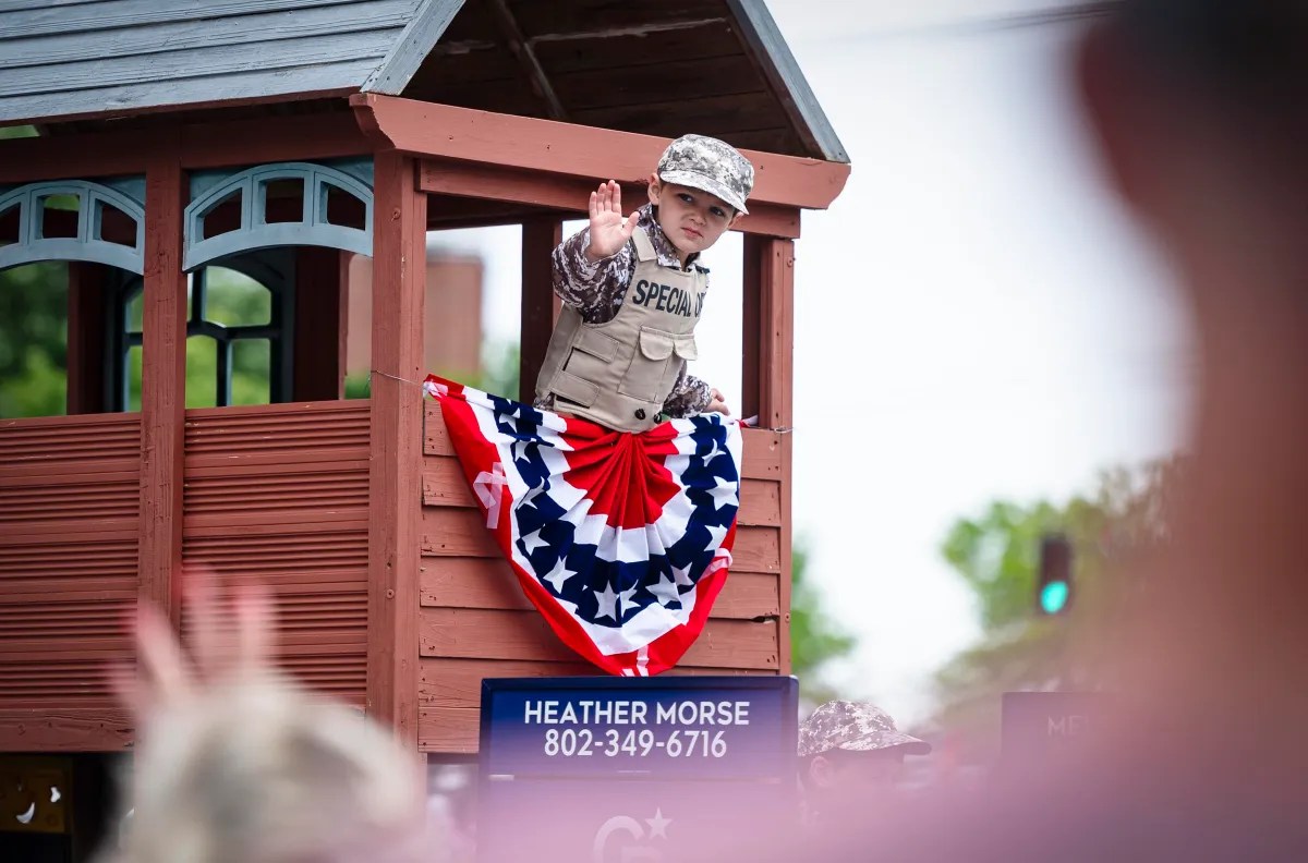 A child in military attire waves from a parade float adorned with patriotic bunting, featuring a sign with a name and phone number.