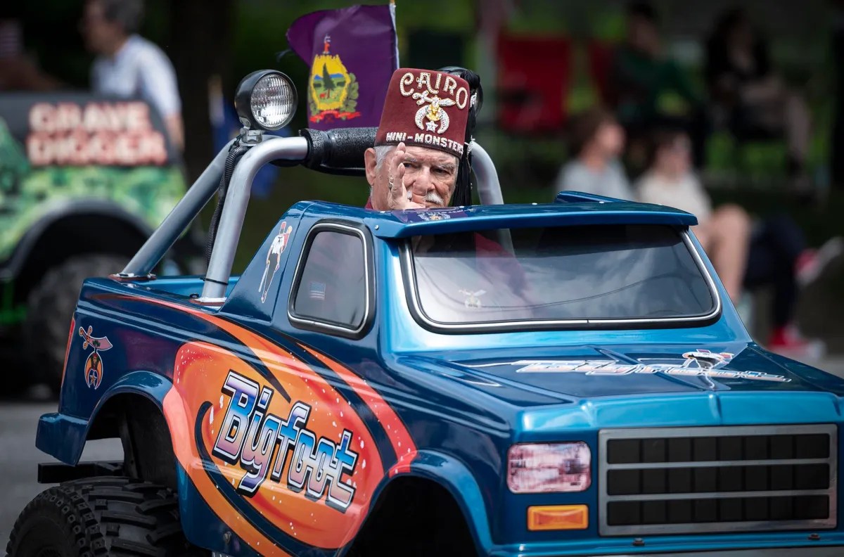 A person wearing a fez hat drives a small, toy-like blue truck labeled "Bigfoot" in a parade. Another person in the background is seated on a float labeled "CRANE EXCAVATOR.