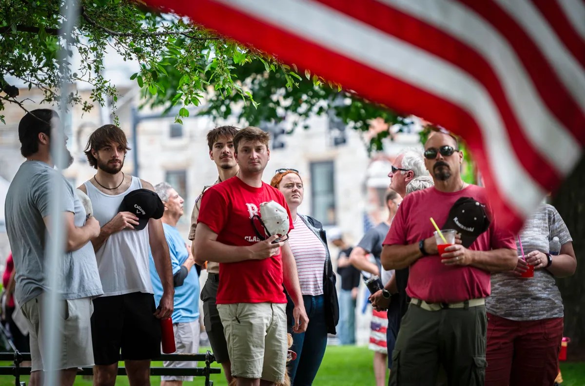 A group of people stands together outdoors, some holding their hats over their hearts, with a large American flag in the foreground.