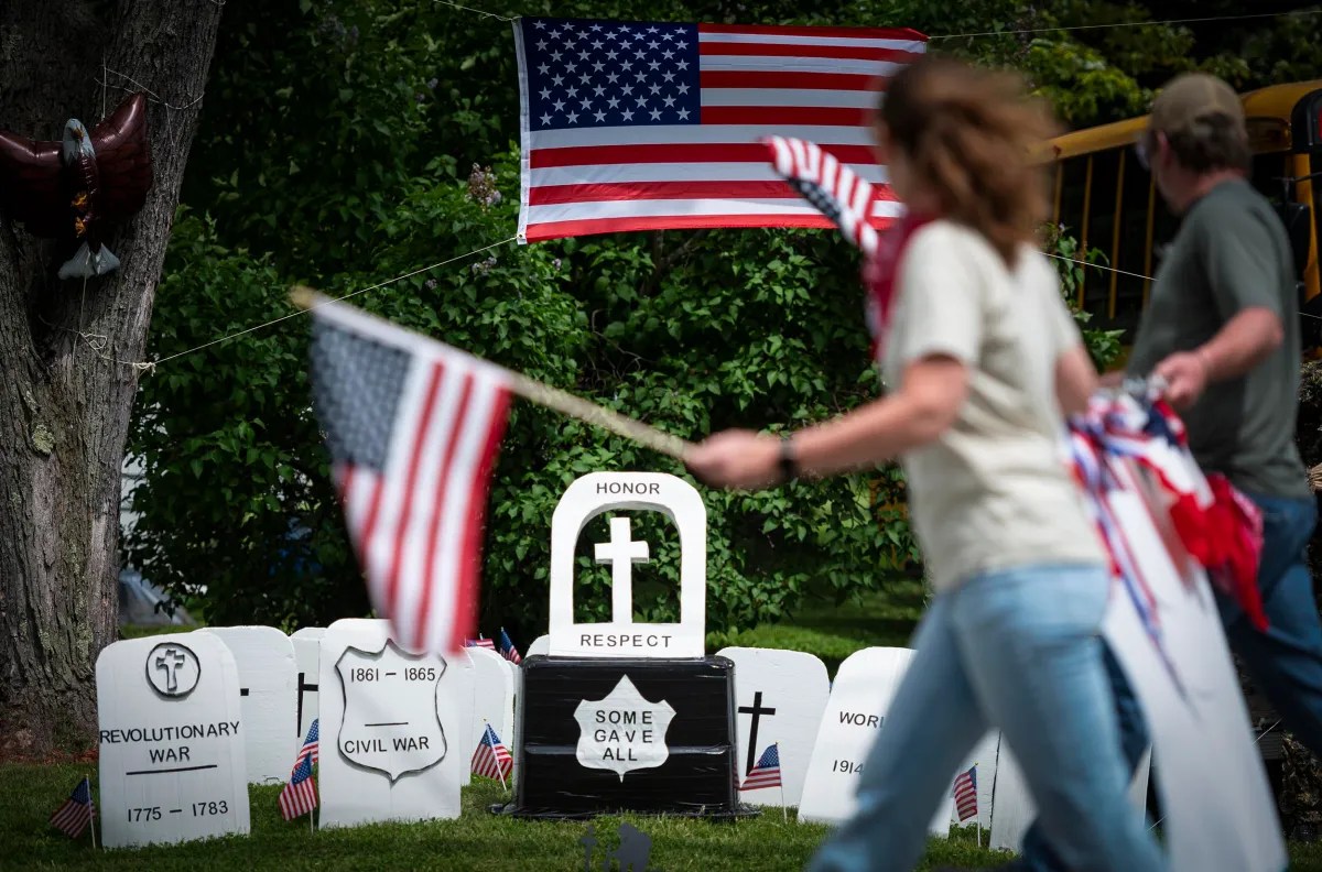 Two people carry American flags in front of a display of headstone replicas honoring fallen soldiers from various wars, with an American flag hung prominently in the background.