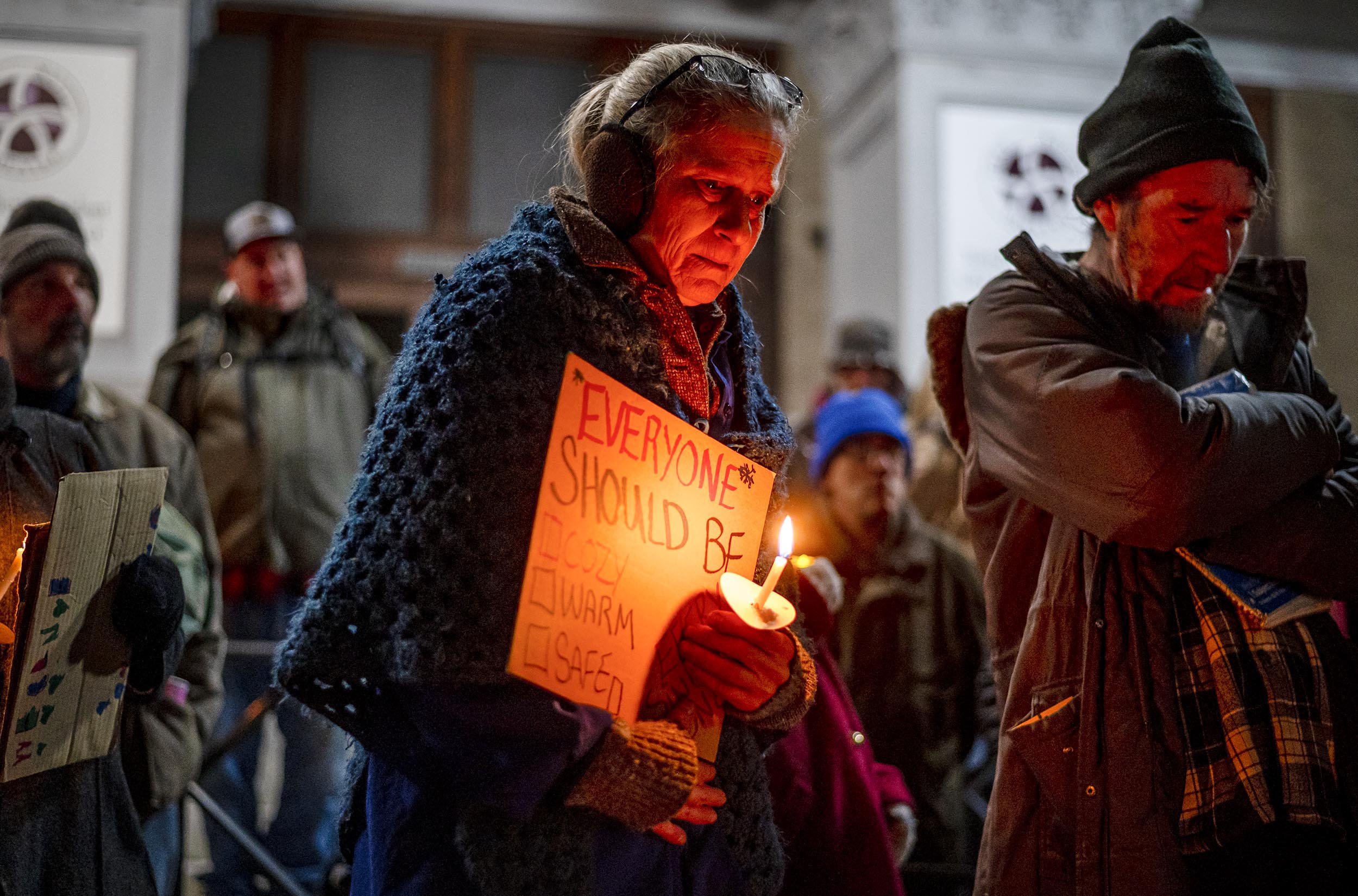 A group of people holding candles, with one person wearing a sign that reads, "Everyone should be warm and safe.