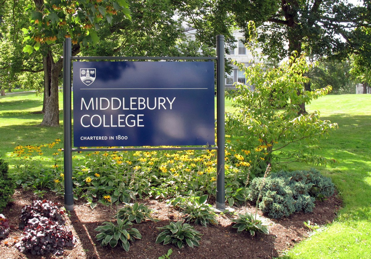 A blue sign reading "Middlebury College, Chartered in 1800" stands in a landscaped area with flowers and greenery, with trees and a building in the background.