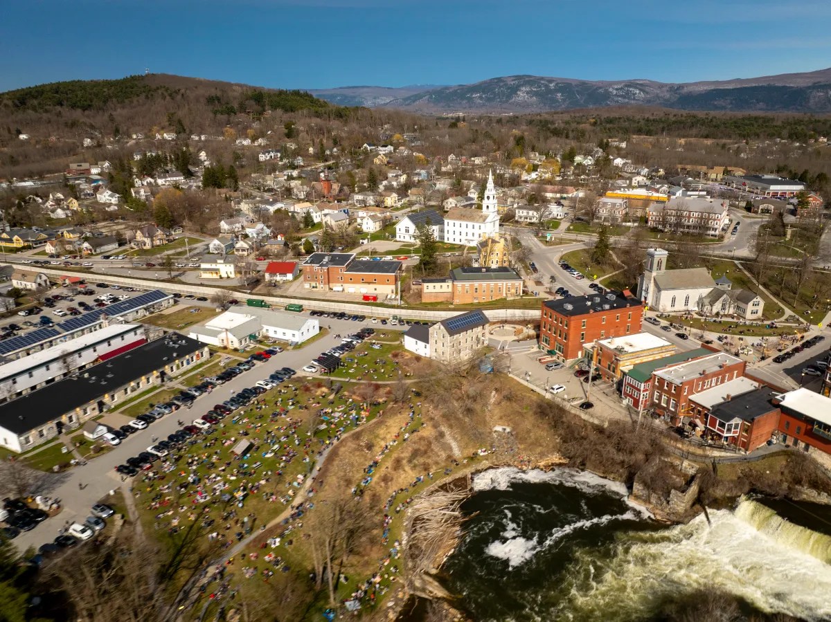 Aerial view of a small town with buildings near a waterfall, showing a community event on a sunny day.