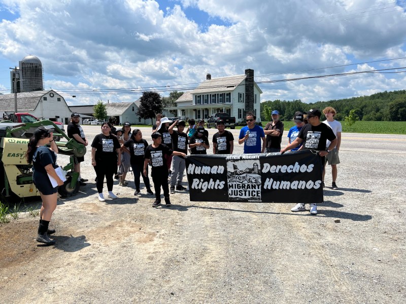 A group of people stands outdoors holding a banner that reads "Human Rights" and "Derechos Humanos." They are gathered on a rural roadside with a farm in the background.