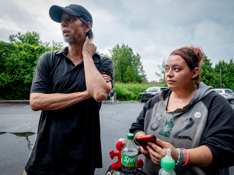 A man in a black cap and shirt stands next to a woman holding a phone; they are outdoors in a parking lot with overcast skies.