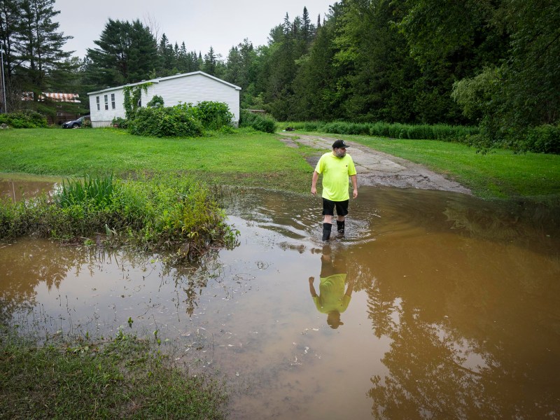A person in a yellow shirt stands in knee-deep floodwater near a rural house surrounded by trees and grass.