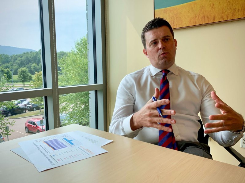 A man in a white shirt and tie sits at a desk explaining something, gesturing with his hands; a printed chart and pen lie on the table beside him.