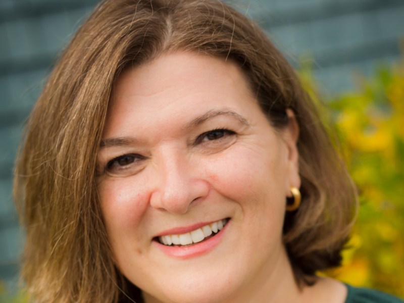 Woman with brown hair and gold hoop earrings smiling outdoors, with greenery and a blurred blue background behind her.