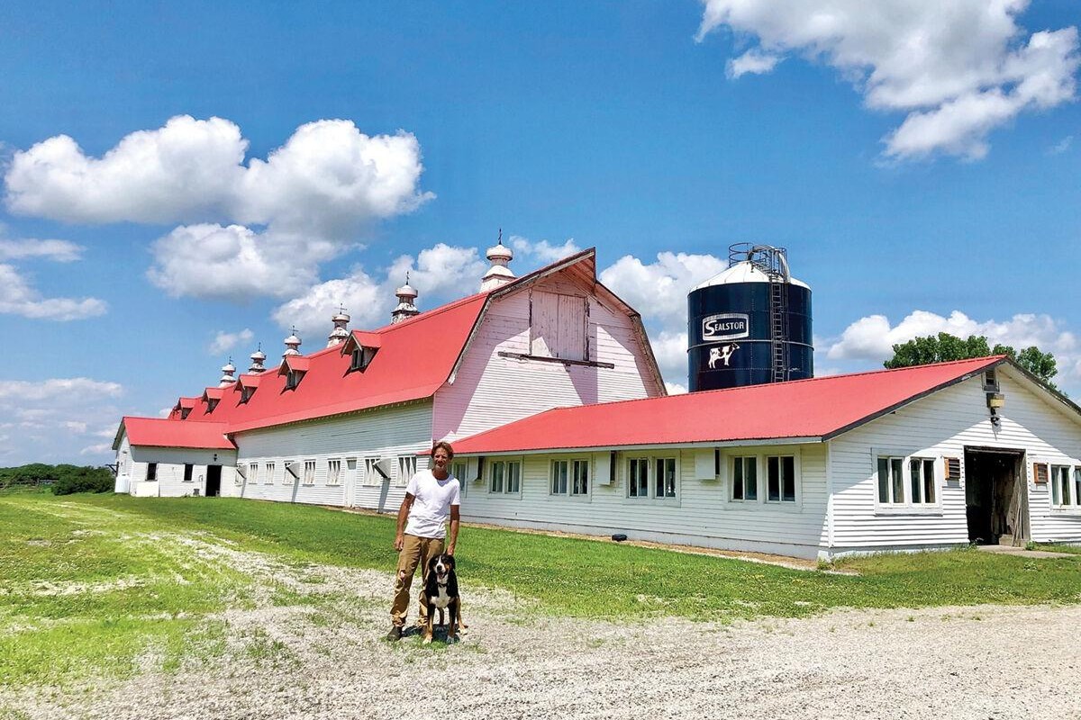 A person stands with a dog in front of a large white barn with a red roof, adjacent to a silo, under a blue sky with scattered clouds.