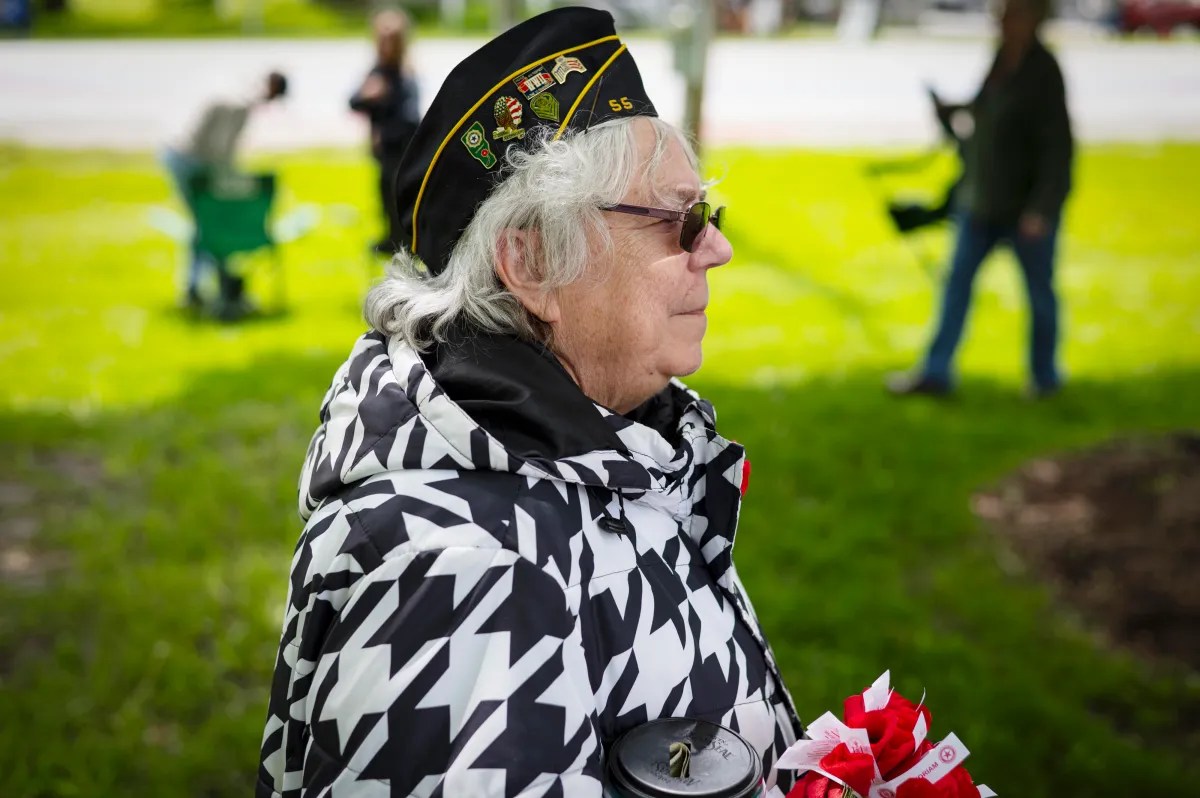 A person in a patterned coat and veteran hat stands outdoors holding flowers, with several people in the background on a grassy area.