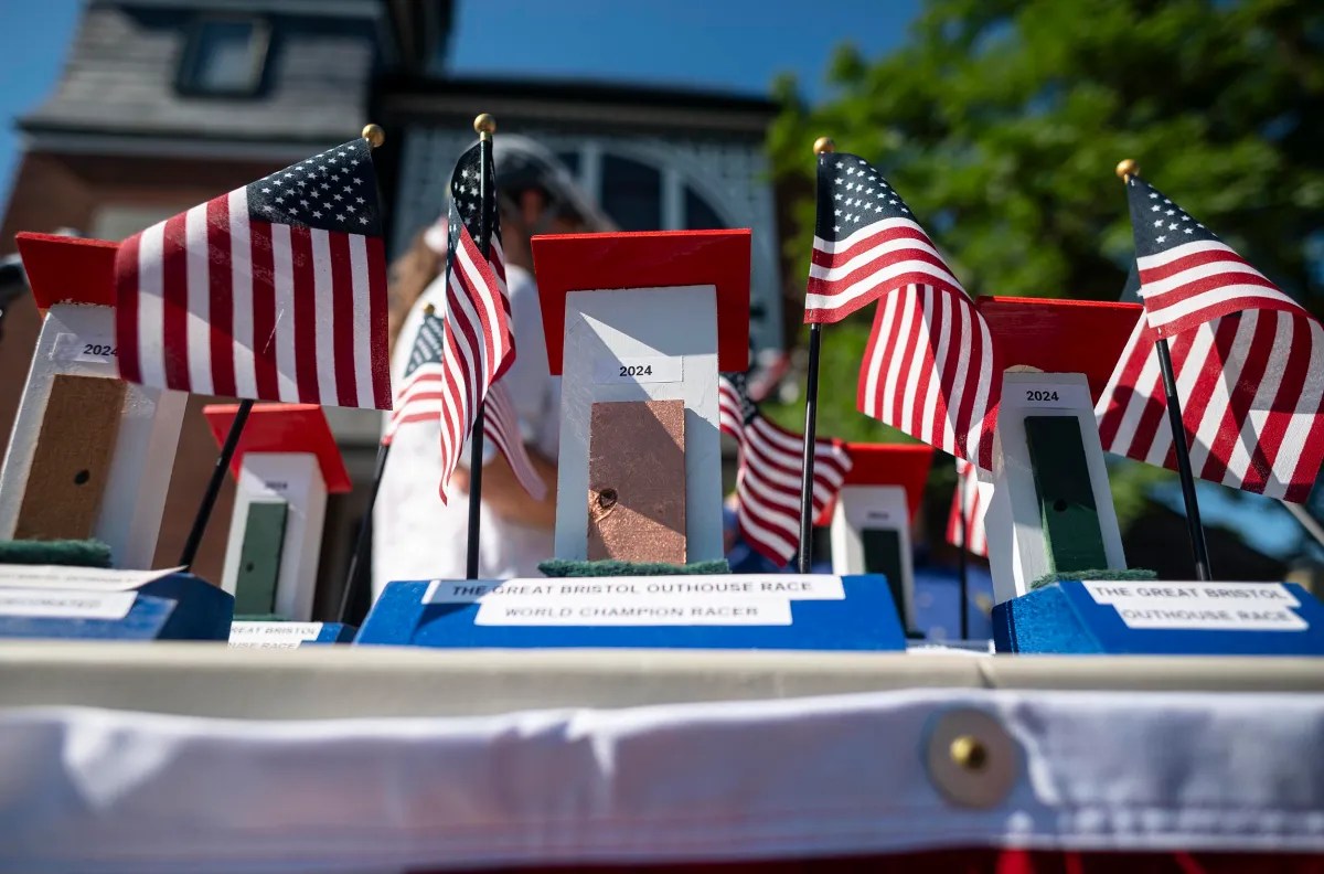 A display table with miniature outhouse models, each topped with an American flag and the year "2024", set outdoors in front of a building.