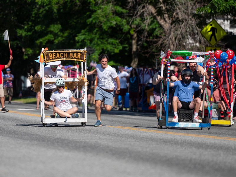 Participants in a soapbox race compete in creatively designed carts on a sunny day, with spectators watching and greenery in the background.